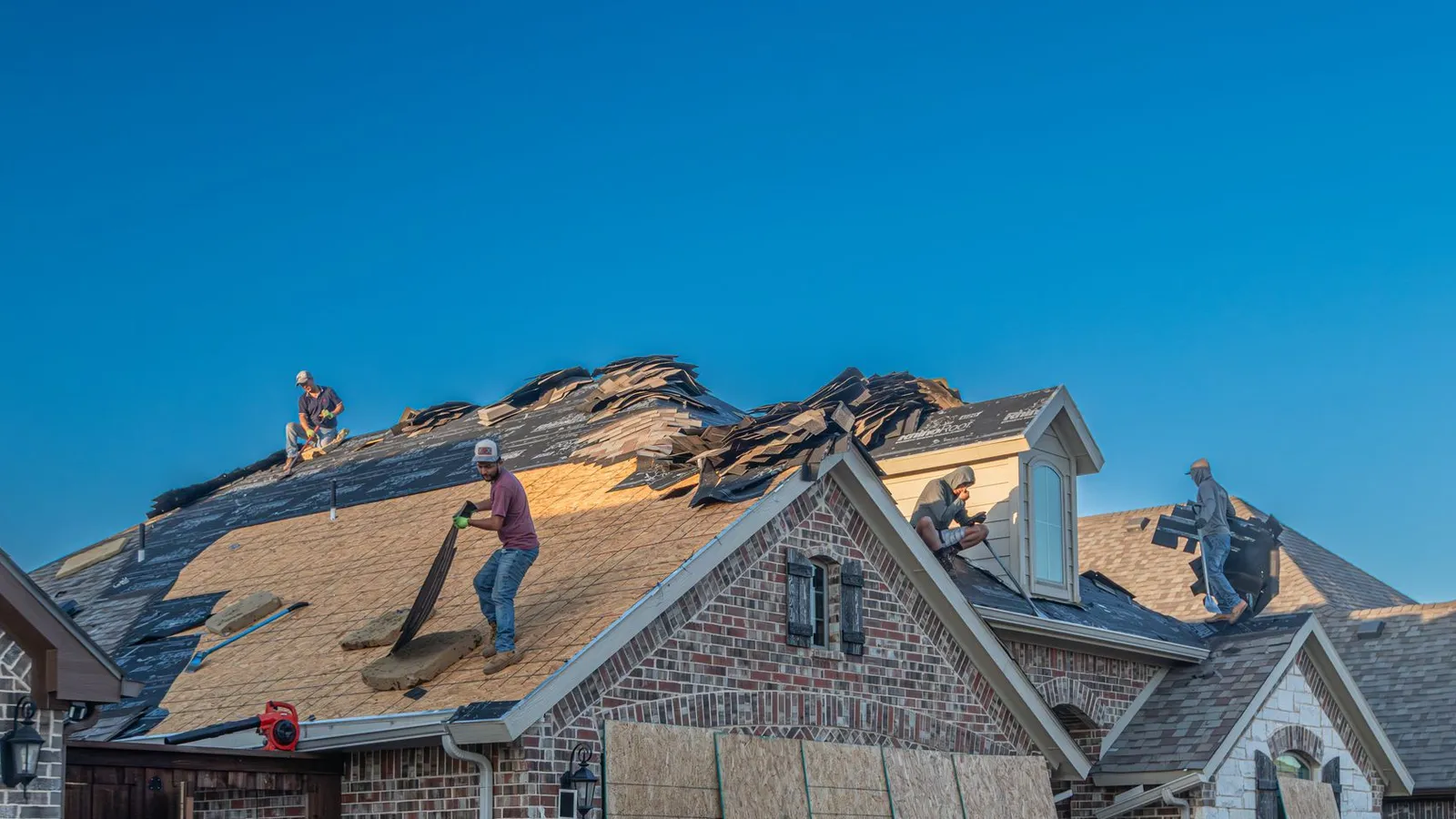 Roofing crew tearing off and replacing shingles on a brick home, work truck parked in the driveway